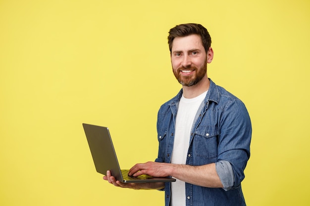 Guapo hombre sonriente con barba sosteniendo la computadora portátil y mirando a la cámara sobre fondo amarillo