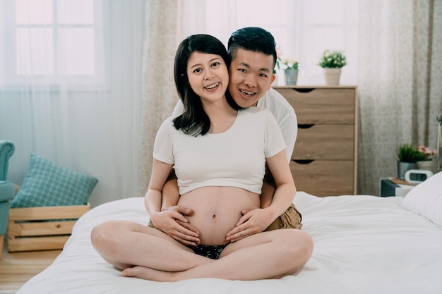 Guapo japonés asiático y hermosa esposa embarazada abrazándose y sonriendo mientras se sienta en la cama cerca de la ventana en el dormitorio de su casa. feliz pareja joven frente a la cámara sonriendo. futuro padre en el día del padre