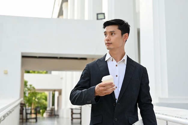 Guapo jefe masculino asiático de pie fuera del edificio con una taza de café en la mano