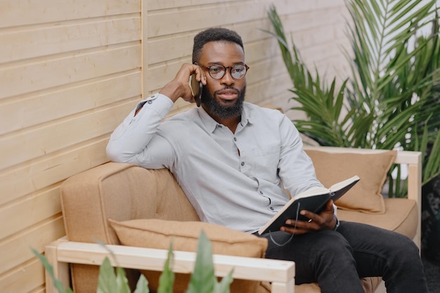 un guapo y joven afroamericano en una oficina o coworking hablando por teléfono y tomando notas en un cuaderno trabajando en una laptop. hombre de negocios afroamericano, comercializador o financiero, moder