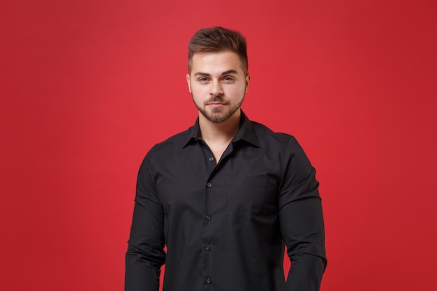 Guapo joven barbudo de 20 años con camisa negra clásica posando aislado en un retrato de estudio de fondo de pared rojo brillante. Emociones sinceras de la gente, concepto de estilo de vida. Simulacros de espacio de copia. Mirando la cámara.