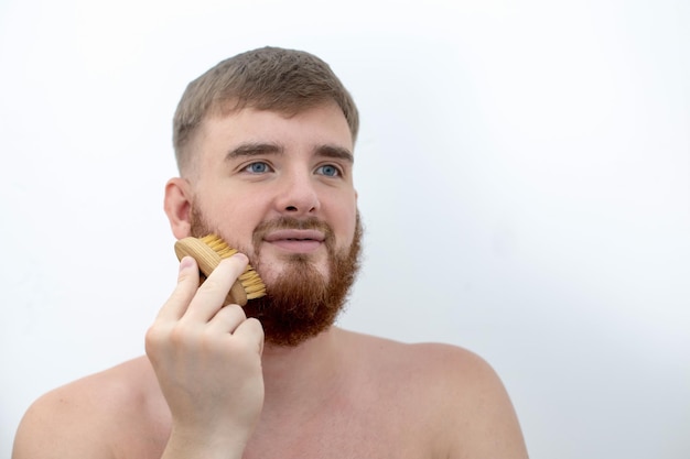 Guapo joven barbudo Hombre brutal europeo con barba está peinando la barba en la cara con un cepillo de peine Accesorio de productos de cuidado personal masculino aislado sobre fondo blanco