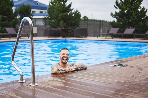 Un guapo joven brutal elegante y elegante chico sonriente posando en la piscina al aire libre