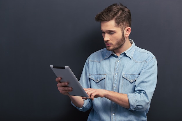 Guapo joven estudiante en camisa de jeans está utilizando una tableta.