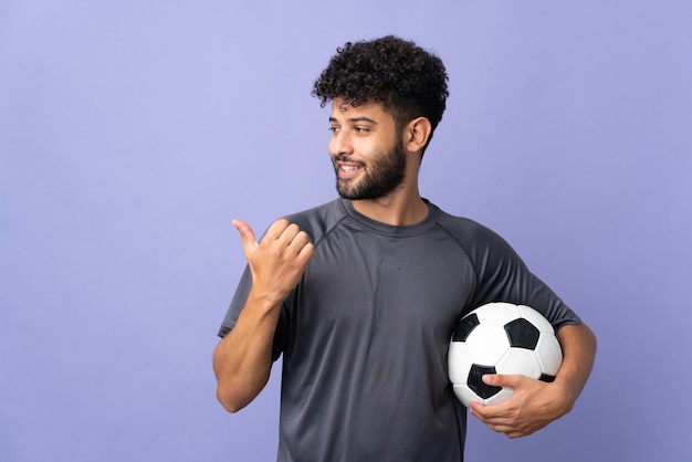 Guapo joven jugador de fútbol marroquí hombre sobre aislado en la pared púrpura apuntando hacia el lado para presentar un producto