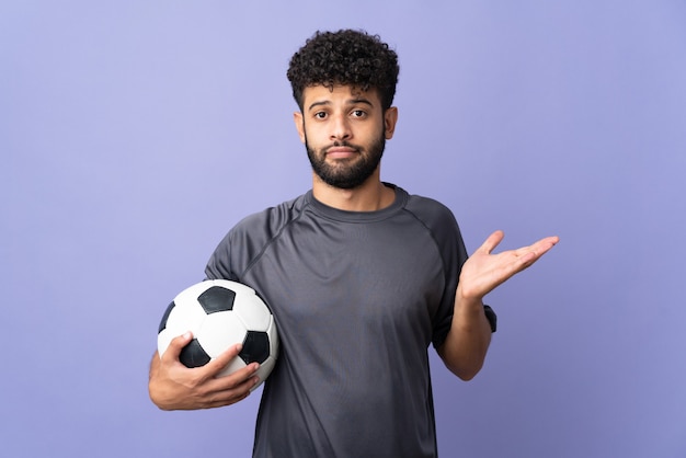 Guapo joven jugador de fútbol marroquí hombre sobre aislado en púrpura con dudas mientras levanta las manos