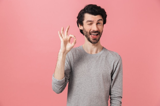 Guapo joven morena barbudo con suéter de pie sobre rosa, mostrando ok