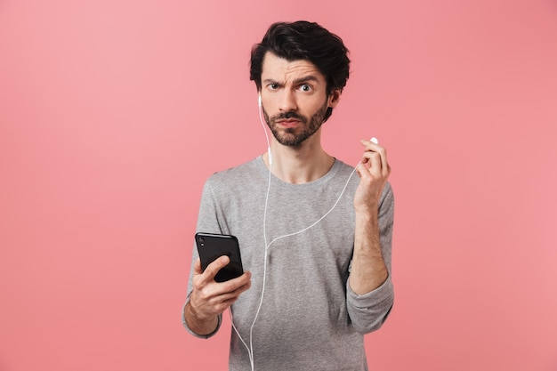 Guapo joven morena barbudo vistiendo un suéter de pie sobre rosa, escuchando música con auriculares, sosteniendo el teléfono móvil