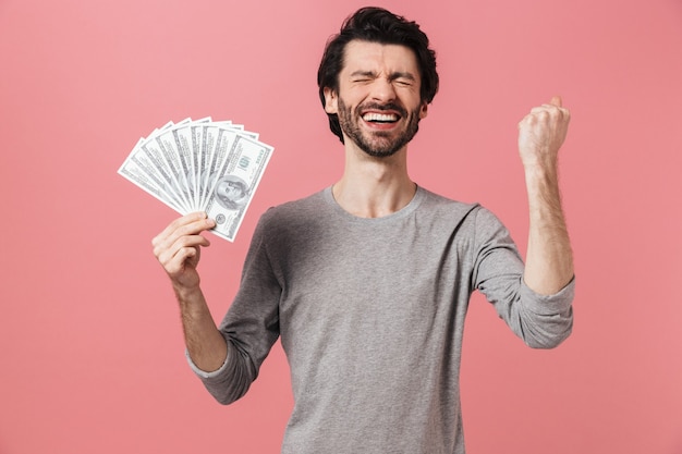 Guapo joven morena barbudo vistiendo suéter de pie sobre rosa, mostrando billetes de dinero