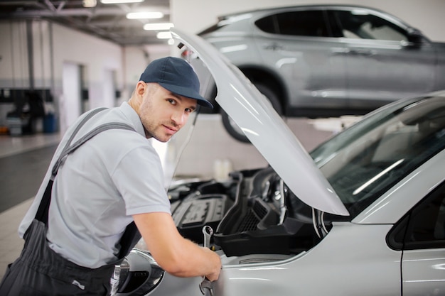 Guapo joven trabajador se ve en la cámara. Hace reparaciones en la parte delantera de la carrocería. Guy usa gorra y uniforme.