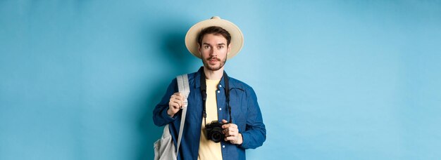 Guapo joven turista en verano sombrero mochilero y viajar con cámara de fotos de pie en blu