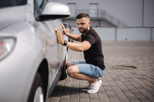 Foto guapo limpia un coche con un trapo en una sala de exposición en un autoservicio de lavado de autos gris