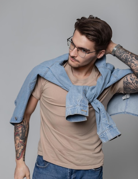 Guapo modelo de estudiante masculino con gafas y peinado en una camiseta simulada de moda con camisa de jeans en el estudio contra un fondo gris