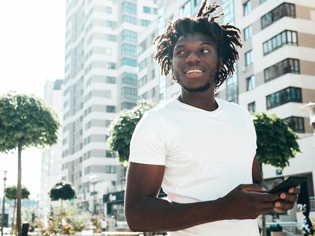 Guapo modelo hipsterHombre africano sin afeitar vestido con camiseta blanca de veranoHombre de moda con peinado de rastas Posando en la calleMirando la pantalla del teléfono inteligente usando aplicaciones de teléfonos celulares