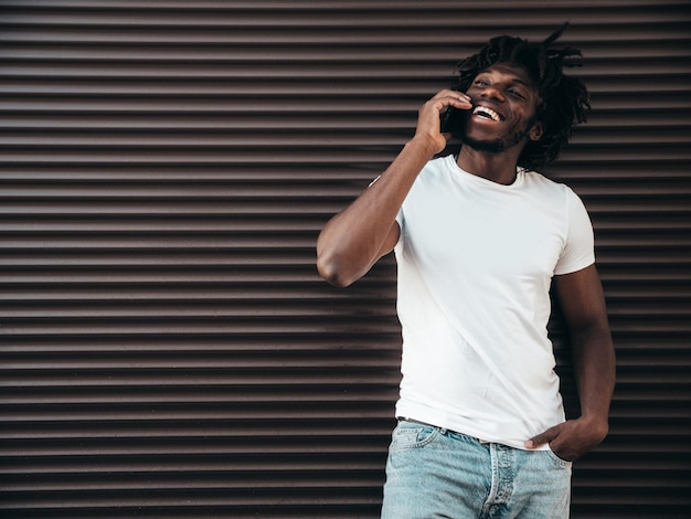 Guapo modelo hipsterHombre africano sin afeitar vestido con camiseta blanca de veranoHombre de moda con peinado de rastas Posando en la calleUsando tiene teléfono inteligente Hablando con amigos en el teléfono celular