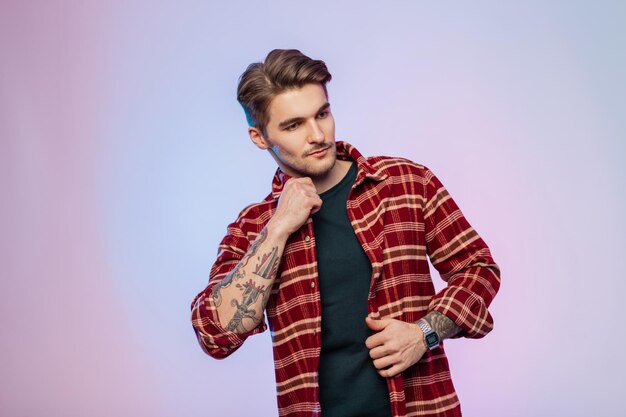 Guapo modelo de hombre hipster con peinado en ropa casual elegante con camisa vintage en estudio con luces creativas coloridas