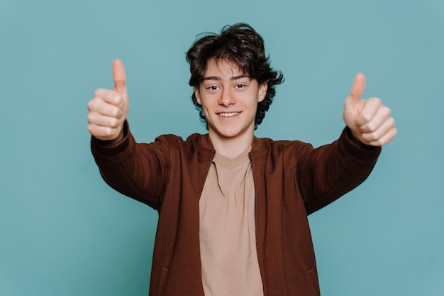 Guapo moreno español con chaqueta marrón y camiseta beige sonríe ampliamente muestra el gesto del pulgar hacia arriba con ambas manos mira la cámara se para contra el fondo turquesa del estudio El tipo alegre aprueba la elección