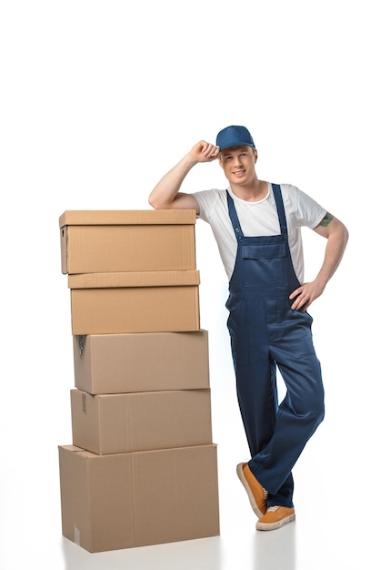 Foto guapo motor en uniforme con cajas de cartón mirando a la cámara mientras ajusta el sombrero aislado en