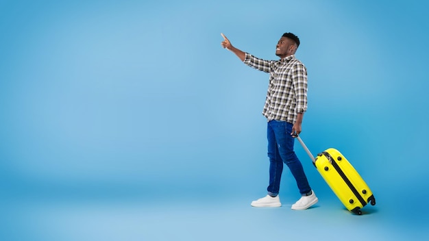 Guapo negro milenario caminando con una maleta brillante apuntando al espacio vacío en el estudio azul