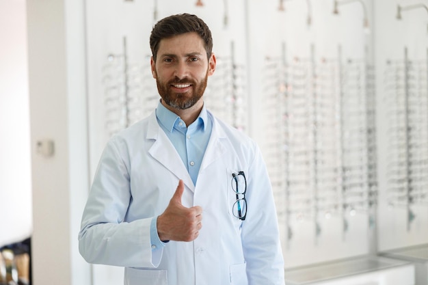 Guapo optometrista sonriente mostrando los pulgares hacia arriba en la tienda de oftalmología