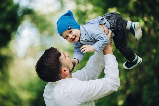 Guapo padre arrojando a un bebé contra el parque de verano Niño pequeño vestido con ropa elegante y papá pasando tiempo juntos sonriendo y divirtiéndose afuera en la naturaleza Concepto de crianza feliz