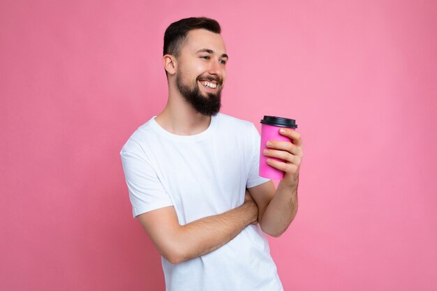 Guapo positivo joven barbudo moreno vestido con camiseta blanca aislada sobre pared rosa backgroung sosteniendo una taza de café de papel bebiendo y mirando hacia el lado