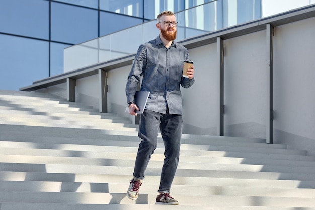 Un guapo rojo independiente con barba camina por la ciudad y bebe café.