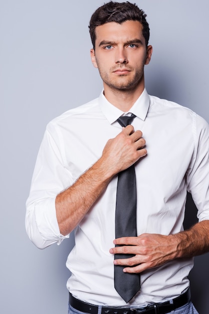 Guapo y seguro de sí mismo. Apuesto joven en ropa formal ajustando su corbata y mirando a la cámara mientras está de pie contra el fondo gris