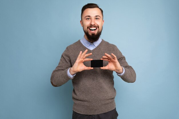 Guapo sonriente morena feliz varón barbudo vistiendo un suéter gris y camisa azul aislado en la pared de fondo con tarjeta de crédito mirando a la cámara