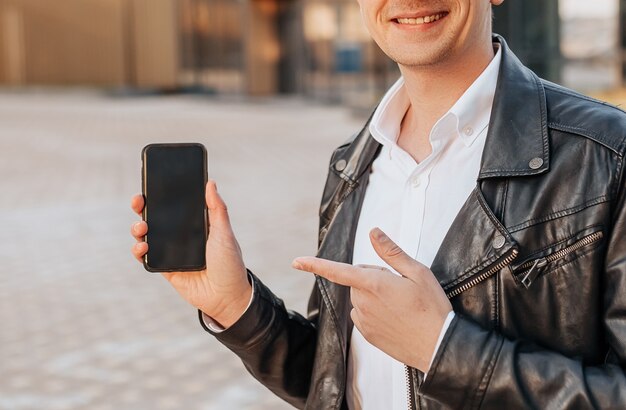 Guapo con un teléfono inteligente en la calle de una gran ciudad. El empresario señala con el dedo a la pantalla del teléfono sobre fondo urbano