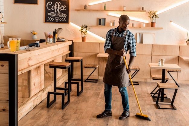 Guapo trabajador afroamericano barriendo con escoba en cafetería