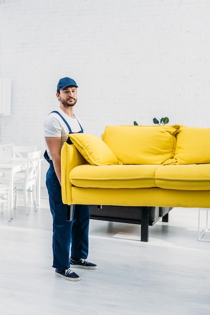 un guapo transportista en uniforme que transporta el sofá en el apartamento