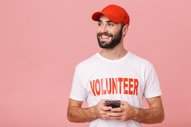 guapo, voluntario, llevando, uniforme, camiseta, sonriente, y, tenencia, teléfono celular, aislado