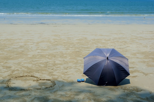 Foto guarda-chuva cinza e chinelos azuis com forma de coração na praia de areia em dia de sol