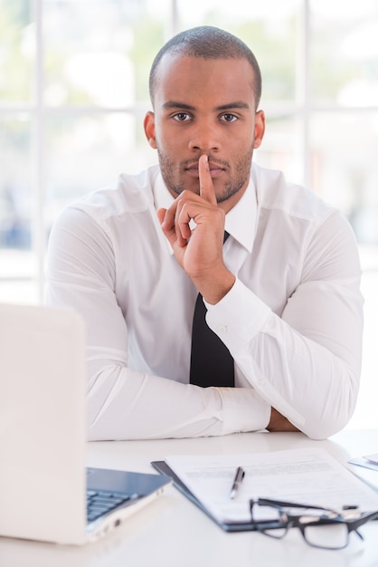 ¡Guarde mi secreto! Apuesto joven africano en camisa y corbata sosteniendo el dedo en los labios