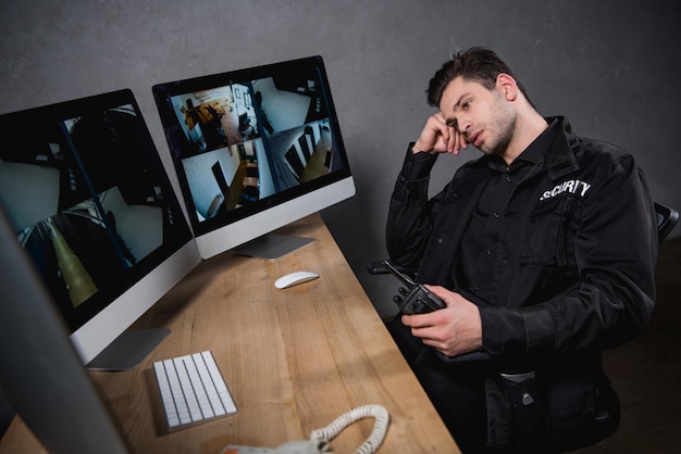 guardia aburrido en uniforme sosteniendo walkietalkie y mirando el monitor de la computadora