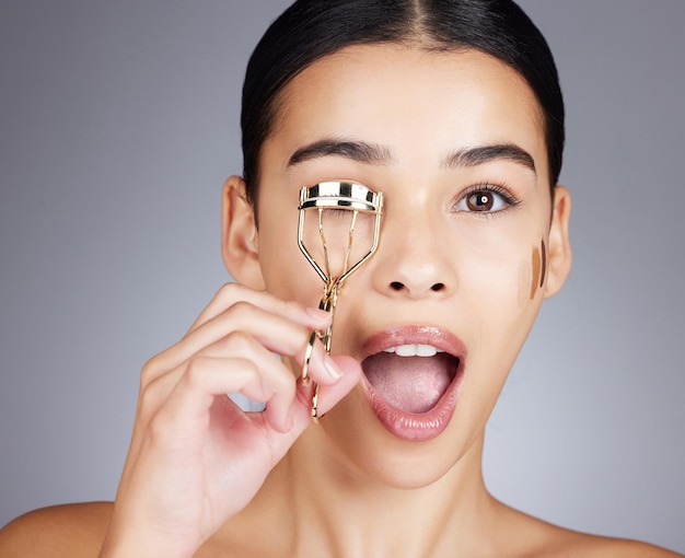 Guau cara y mujer con rizador de pestañas en estudio aislado en un fondo gris Sorpresa de maquillaje de retrato y cosméticos de modelo femenino con herramienta o producto para rizar pestañas por estética de belleza