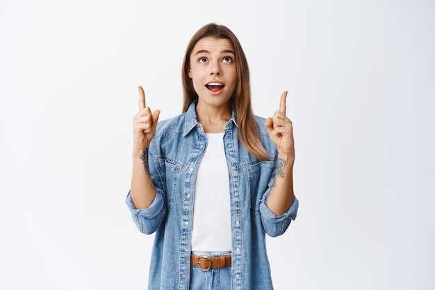 Guau, mira Chica rubia sorprendida mirando el trato promocional mirando y señalando con el dedo hacia arriba en el anuncio superior que muestra una oferta genial de fondo blanco