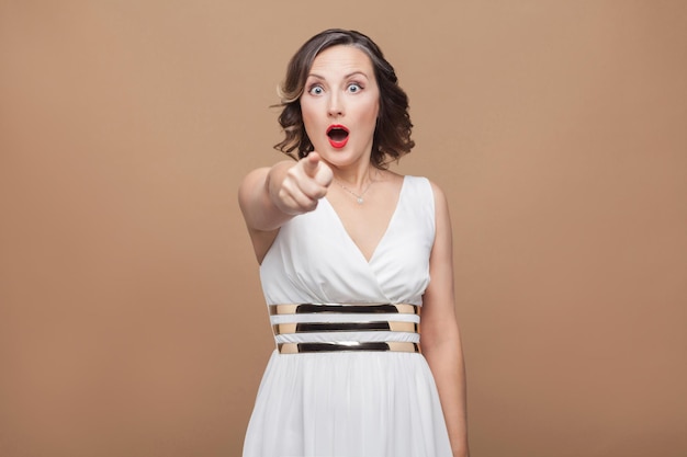 ¡Guau! Mujer adulta sorprendida que señala el dedo a la cámara. Mujer de expresión emocional en vestido blanco, labios rojos y peinado rizado oscuro. Foto de estudio, interior, aislado sobre fondo beige o marrón claro