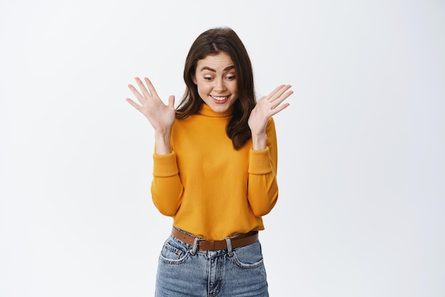 Guau, súper emocionada chica morena saltando y aplaudiendo de la emoción y la alegría mirando hacia abajo y sonriendo, ve noticias increíbles que reaccionan a un fondo blanco sorprendentemente sorprendente