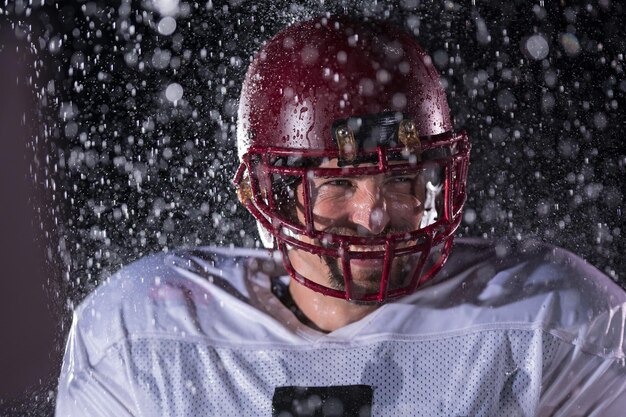 El guerrero atleta de fútbol americano parado en un campo sostiene su casco y está listo para jugar. Jugador preparándose para correr, atacar y anotar touchdown. Noche lluviosa con destello de lente dramático y gotas de lluvia. Alto