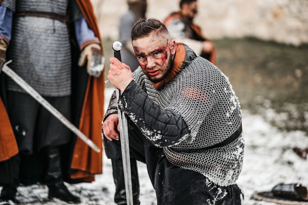 Un guerrero medieval con armadura de cota de malla y manto se arrodilla frente a los soldados que han muerto en la batalla.