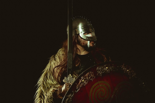 Guerrero vikingo con casco metálico y pieles de animales. guanteletes de cuero, escudo de madera rojo con adornos dorados y espada de acero