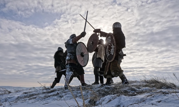 Los guerreros de los vikingos luchan durante el ataque en invierno. Los vikingos luchan con espadas, escudos y cascos en el prado de invierno. Los guerreros de los vikingos luchan contra el cielo nublado con sol