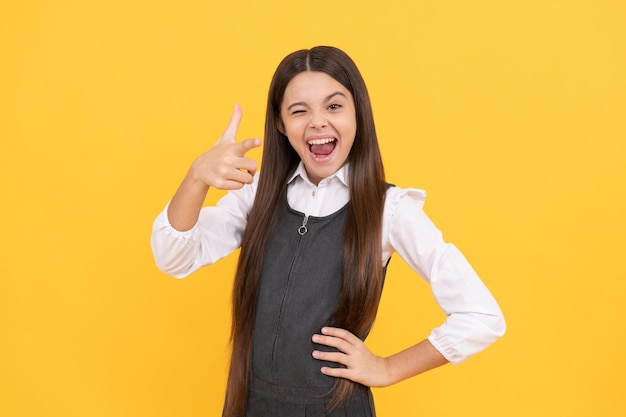 Guiño en edad escolar niña feliz sonrisa apuntando con el dedo pistola mano gesto fondo amarillo, 1 de septiembre.