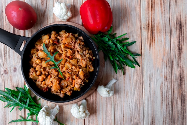 Guisado de carne caseiro com legumes, batata, cebola, cenoura, couve-flor, pimenta com molho de tomate, alho e ervas em uma frigideira de ferro fundido na mesa de madeira. Cama plana rústica. Copie o espaço. Vista do topo.