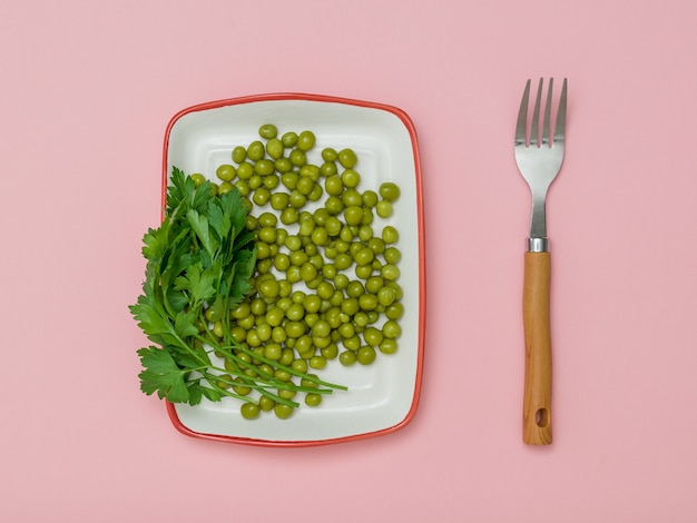 Guisantes enlatados en un plato blanco y un tenedor sobre un fondo rosa. Comida vegetariana dietética.