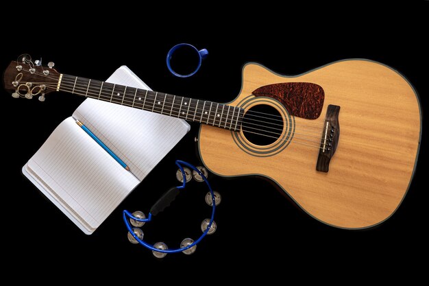 Guitarra acústica, pandeiro, bloco de notas aberto e xícara de café em fundo preto, vista superior, conceito de criatividade musical.