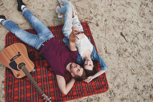 Guitarra acústica perto deles. Jovem casal faz piquenique na praia. Deitado no cobertor de cor vermelha.