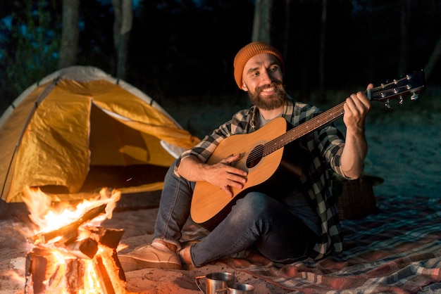 Foto guitarrista acampando en la noche junto a una fogata.